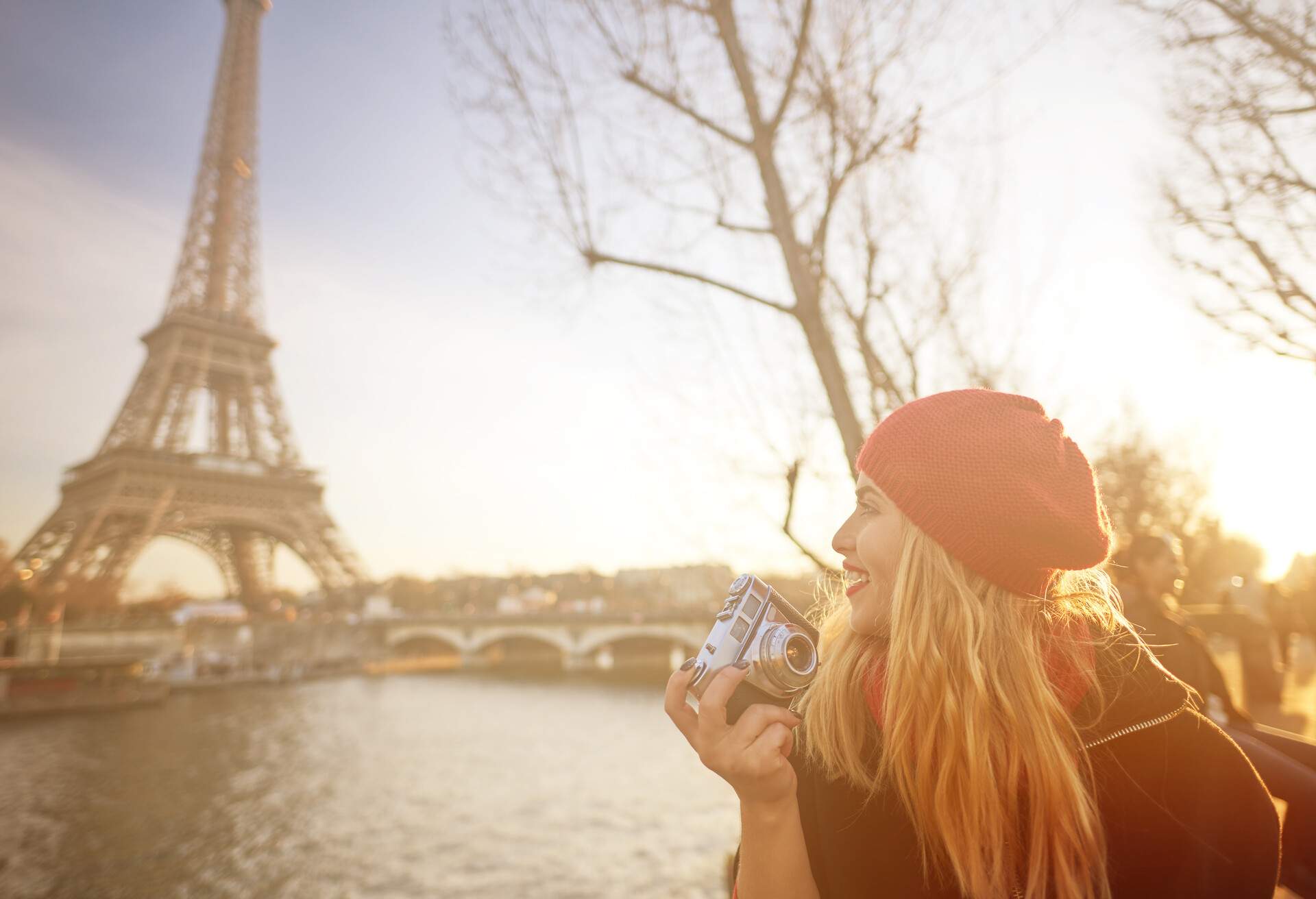 A woman with a camera smiling as she looks towards the Eiffel Tower from across the river.