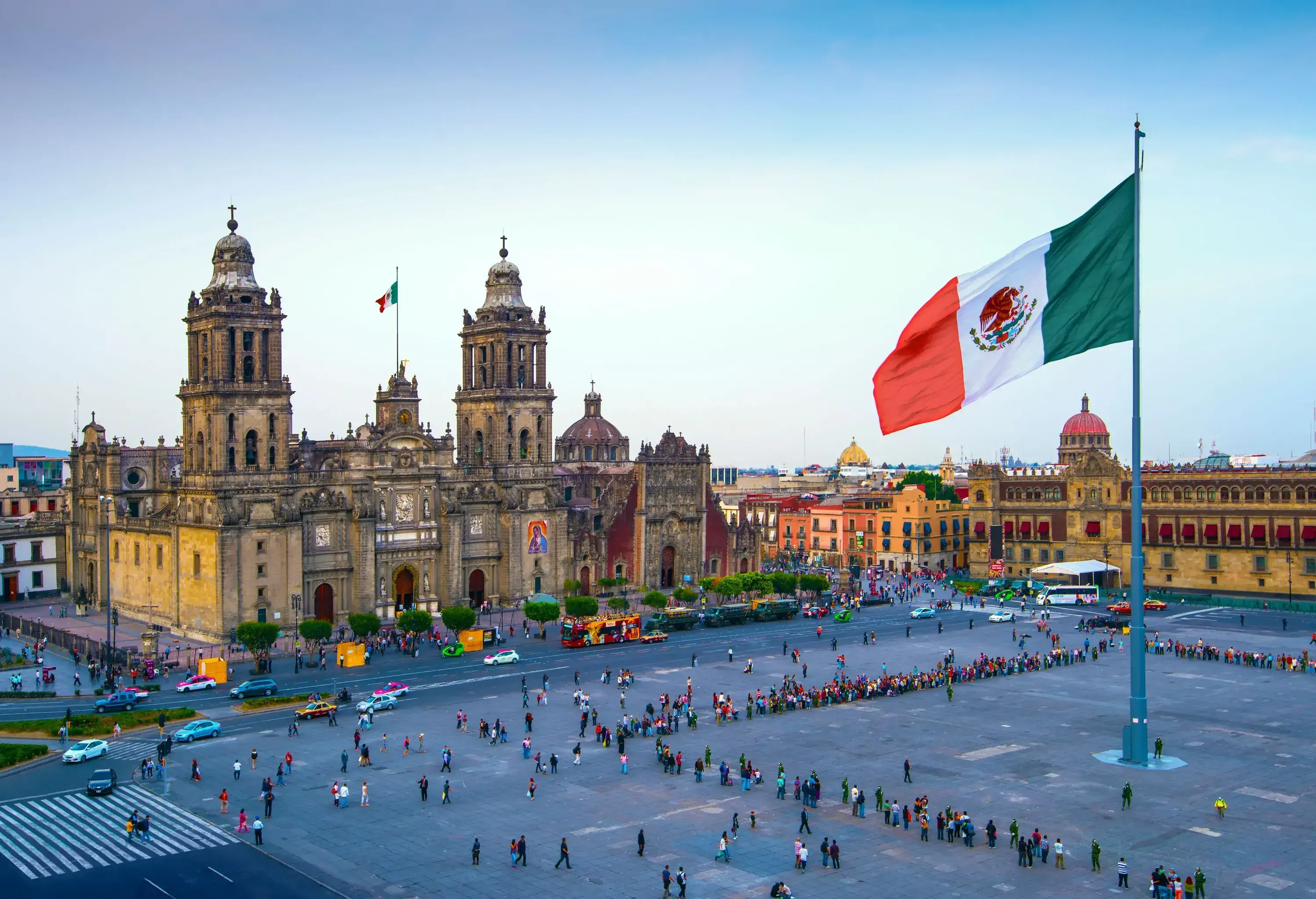 People gather on the square with a Mexican flag in the centre, surrounded by a church and colourful buildings.
