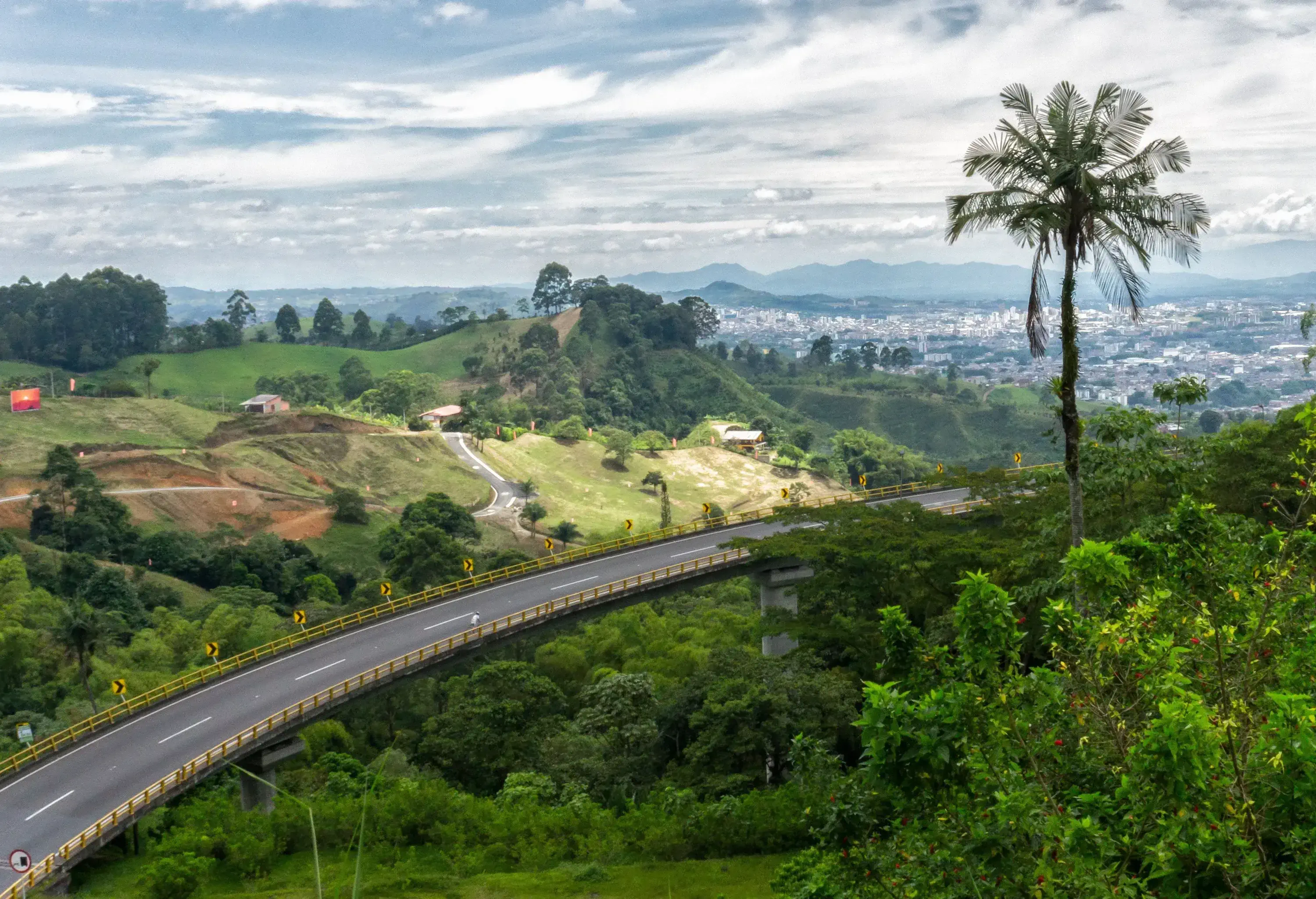A two-lane highway elevated over a hilly terrain densely forested with trees and plants.