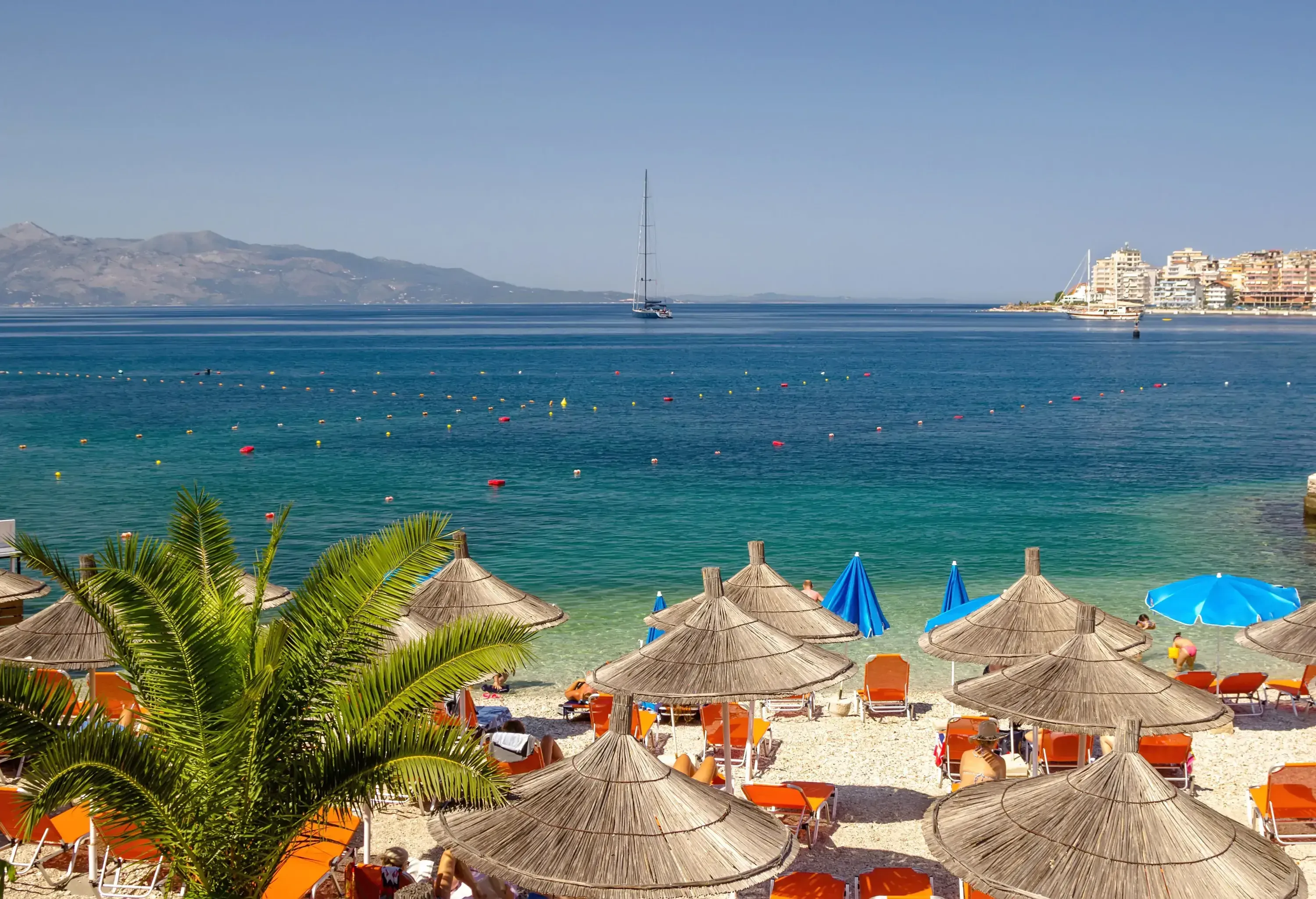 Sun umbrellas and beach chairs set up on a sandy beach, overlooking a tranquil bay with distant mountains and buildings.